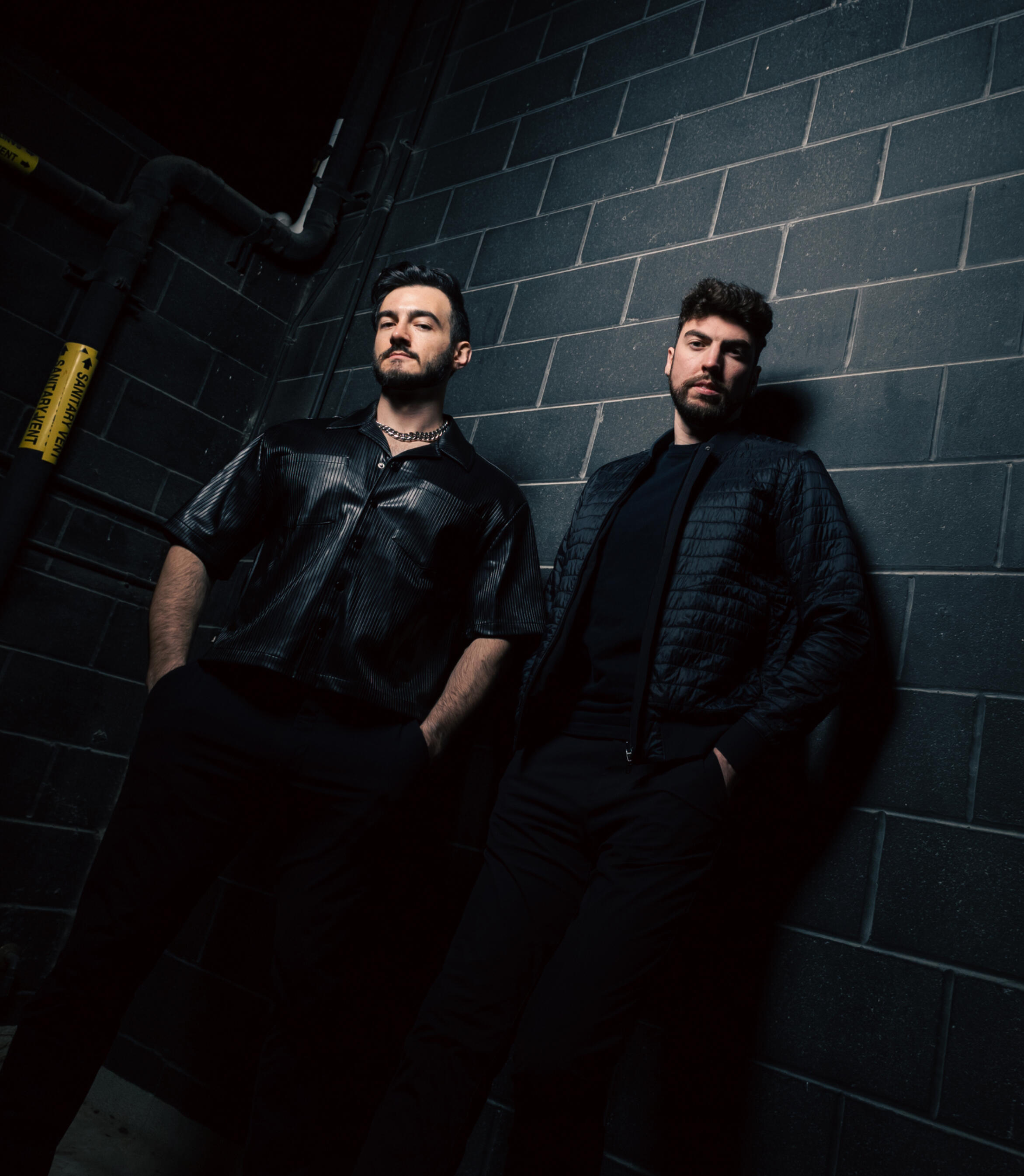 saintstratus-presspic01 Saint Stratus, a DJ duo of two men, wearing all black and standing near a black brick wall